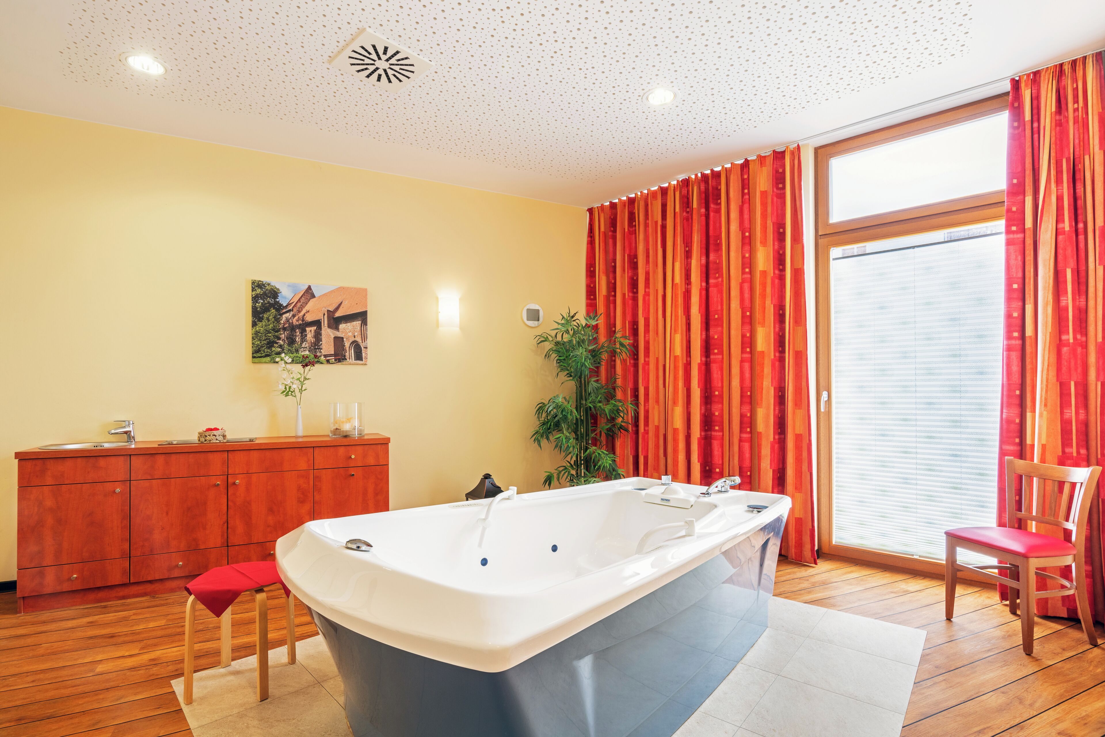 A bathroom with a bathtub, wooden floor, a wooden cabinet, a chair, a plant, and a window with curtains.