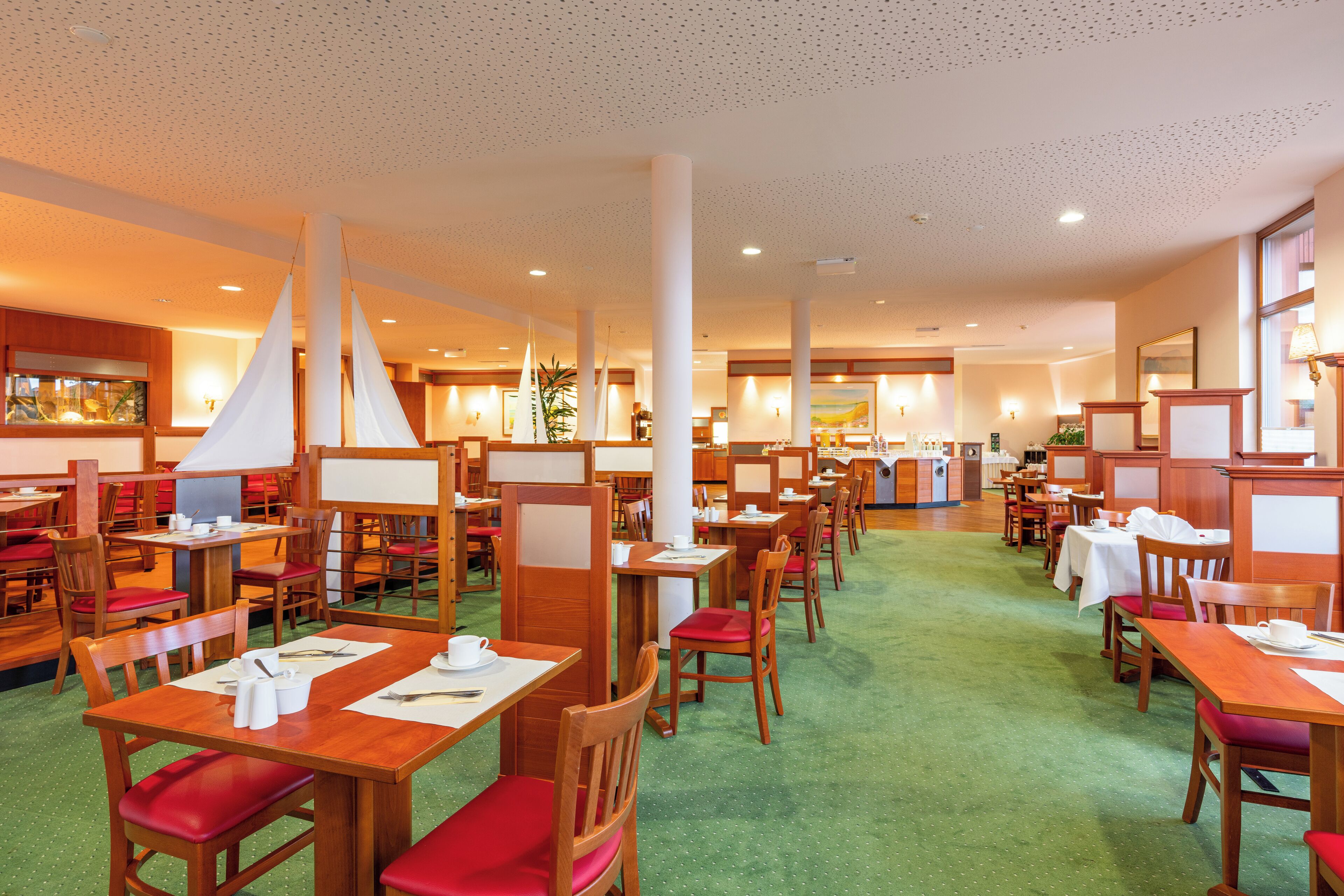 Ein geräumiges Restaurant mit Holztischen und -stühlen, einem grünen Teppichboden und einer hohen Decke mit punktiertem Muster.