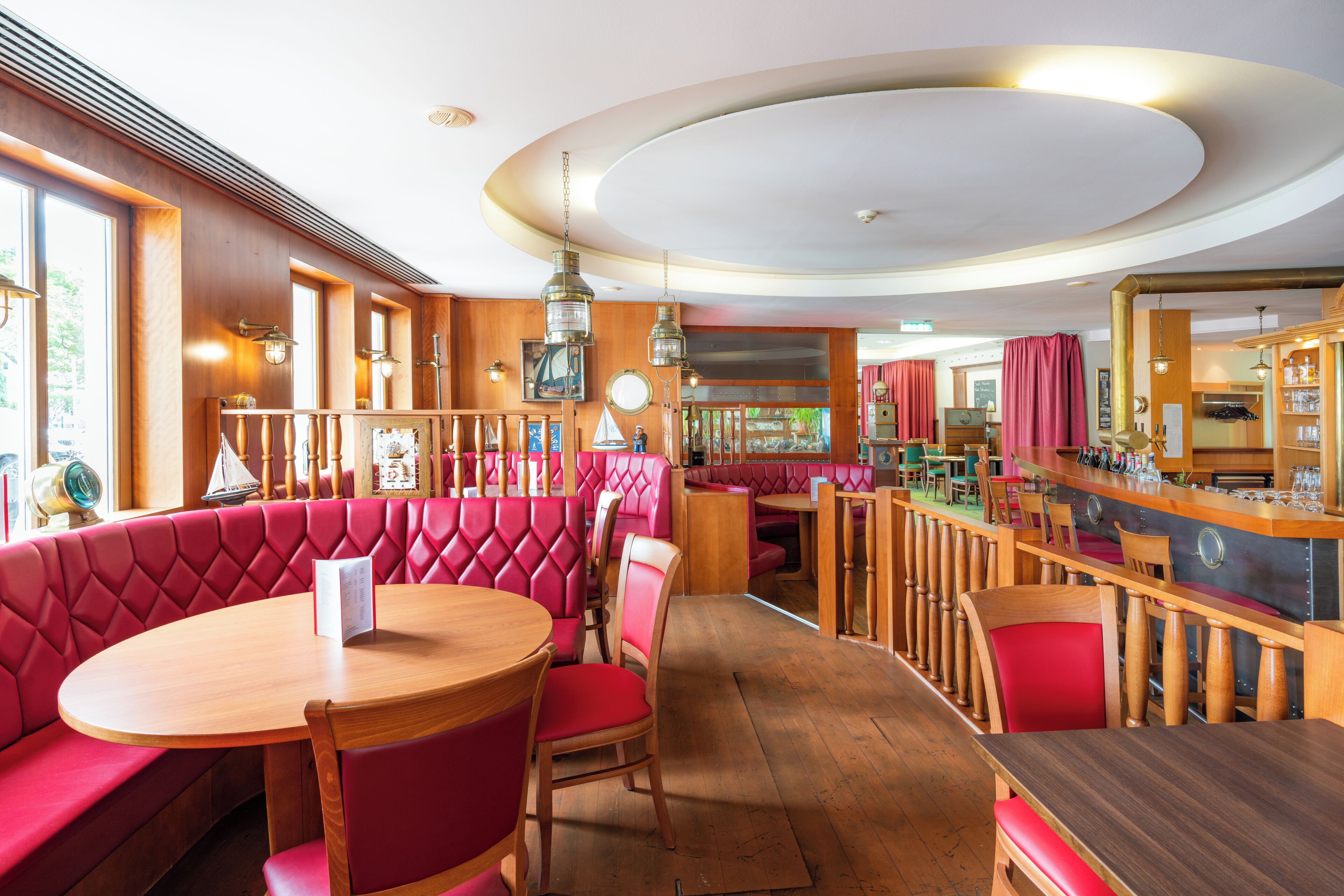 A restaurant with red upholstered booths, wooden tables, and a bar area.