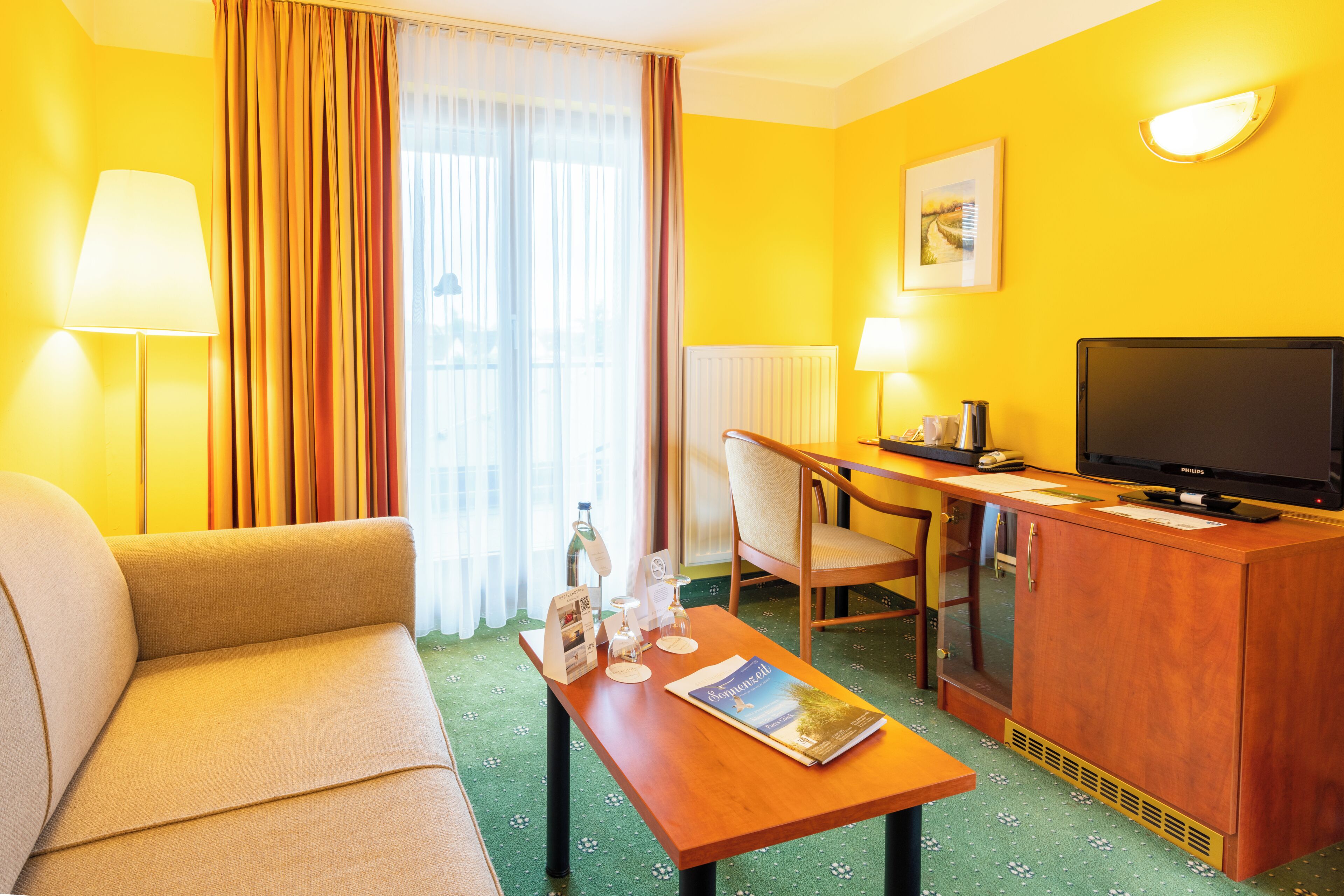 Habitació d'hotel amb sofà, escriptori amb televisió, cadira i taula petita amb una revista.