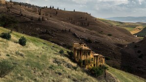 Un bâtiment rustique en pierre, doté d’un toit en pente, entouré de collines douces et d’une végétation clairsemée.