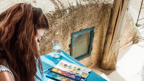 A person drawing at a table with a blue tablecloth, a glass of water, and a watercolor palette. - Beit Al Fannan (Tabaqat Fahl)
