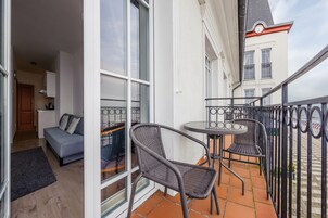 Un balcón con sillas de mimbre y una mesita, un sofá en el interior, y vista a un edificio con un reloj.