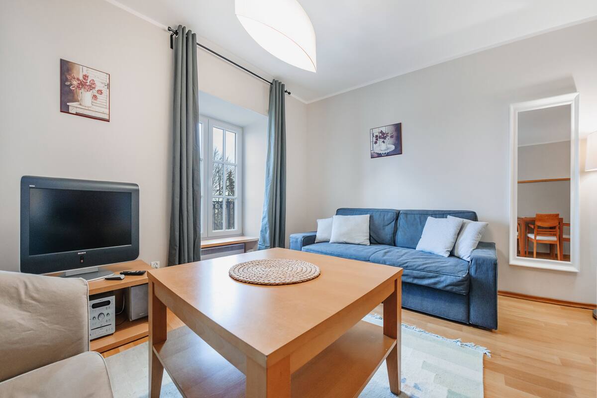 Ein modernes Wohnzimmer mit Flachbildfernseher, Couchtisch aus Holz, blauem Sofa und einem Fenster mit Vorhängen.