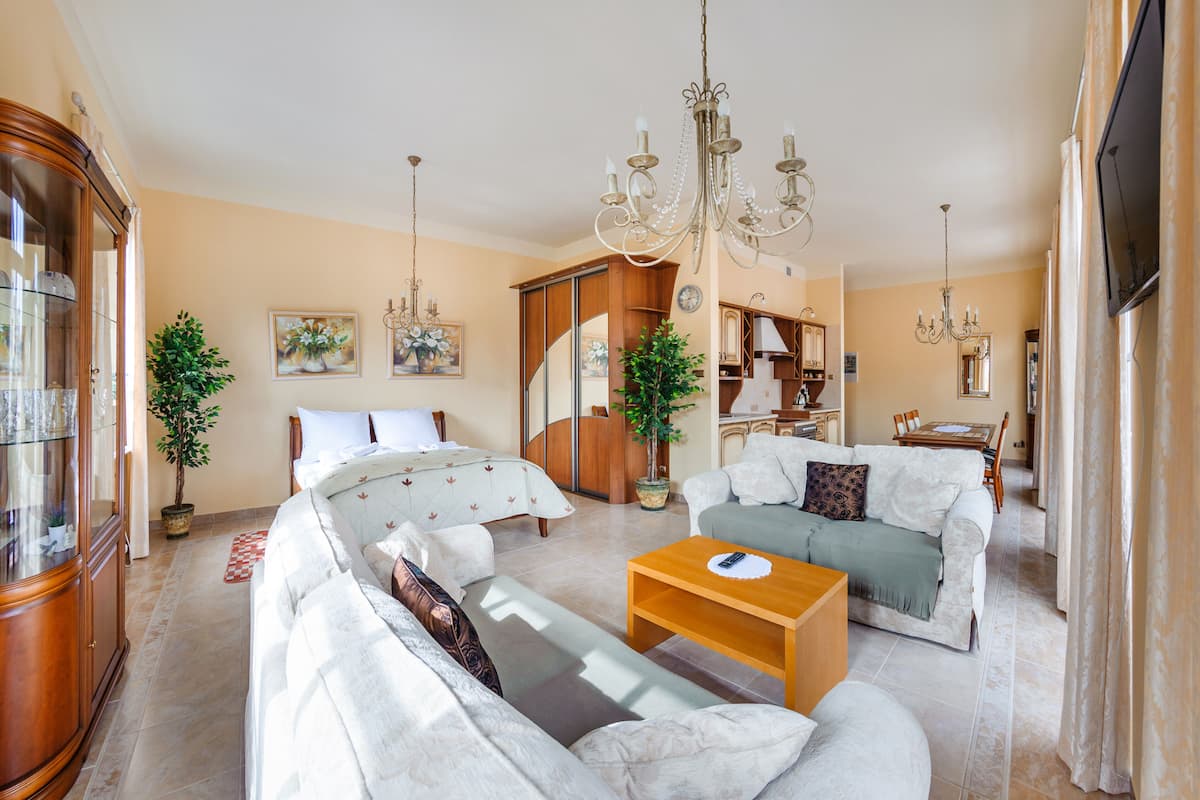 Ein geräumiges Wohnzimmer mit einer Sofagarnitur, einem Holztisch und einer Chandelier. Es gibt einen Essbereich mit Tisch und Stühlen, und im Hintergrund eine Küche.