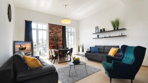 A modern living room with a sofa, armchair, and coffee table. There is a dining area with a table and chairs. The room has a brick wall, large windows, and shelves with plants. - Apartamenty Sun&Snow Zachód Slonca (Mielno)