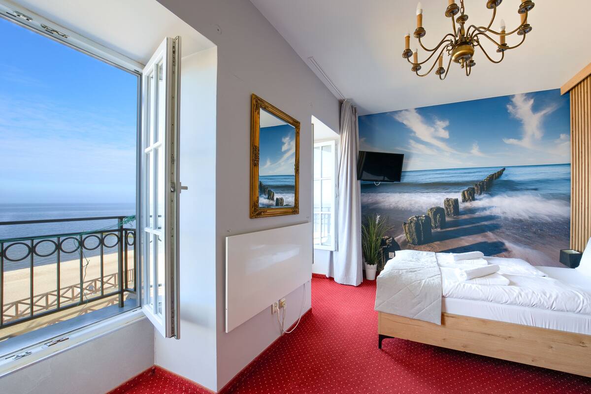 Ein Schlafzimmer mit großem Fenster und Meerblick, einem Bett mit weißer Bettwäsche und einem Wandgemälde mit Strandmotiv.