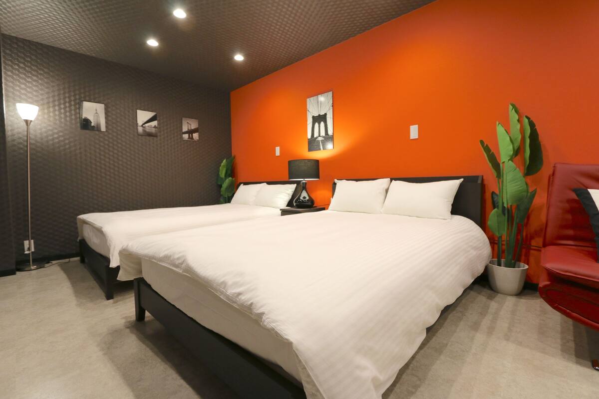 Quarto de hotel com duas camas, uma cadeira vermelha, um abajur e uma planta em vaso.
