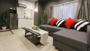 A modern living room with a grey sofa, red and black striped pillows, a wooden coffee table with a decorative runner, a kitchen area with a built-in oven and microwave, and green plants. - Elizabeth Garden Shinmachi (Osaka)
