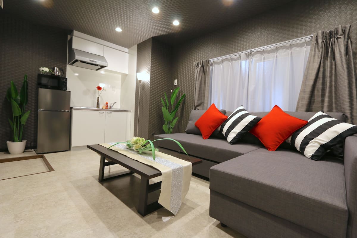 Uma sala de estar moderna com um sofá cinzento, almofadas listradas em vermelho e preto, uma mesa de centro de madeira com um tapete decorativo, uma área de cozinha com forno embutido e micro-ondas, e plantas verdes.