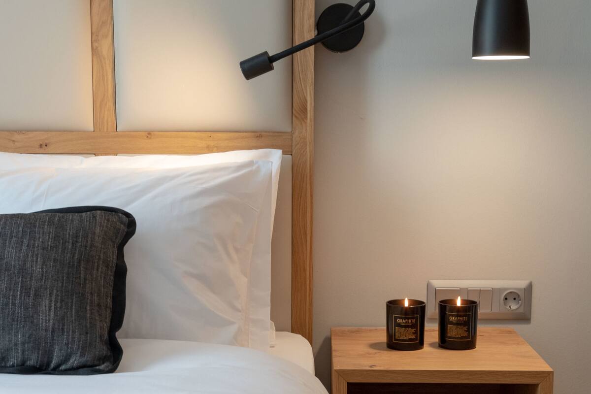 Una cama bien hecha con un almohadón gris, una lámpara de noche y dos velas sobre una mesita de madera.