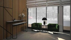 Un interior moderno con un panel de madera, un banco, dos sillones verdes, una mesita con una lámpara y grandes ventanas con vistas a la calle.