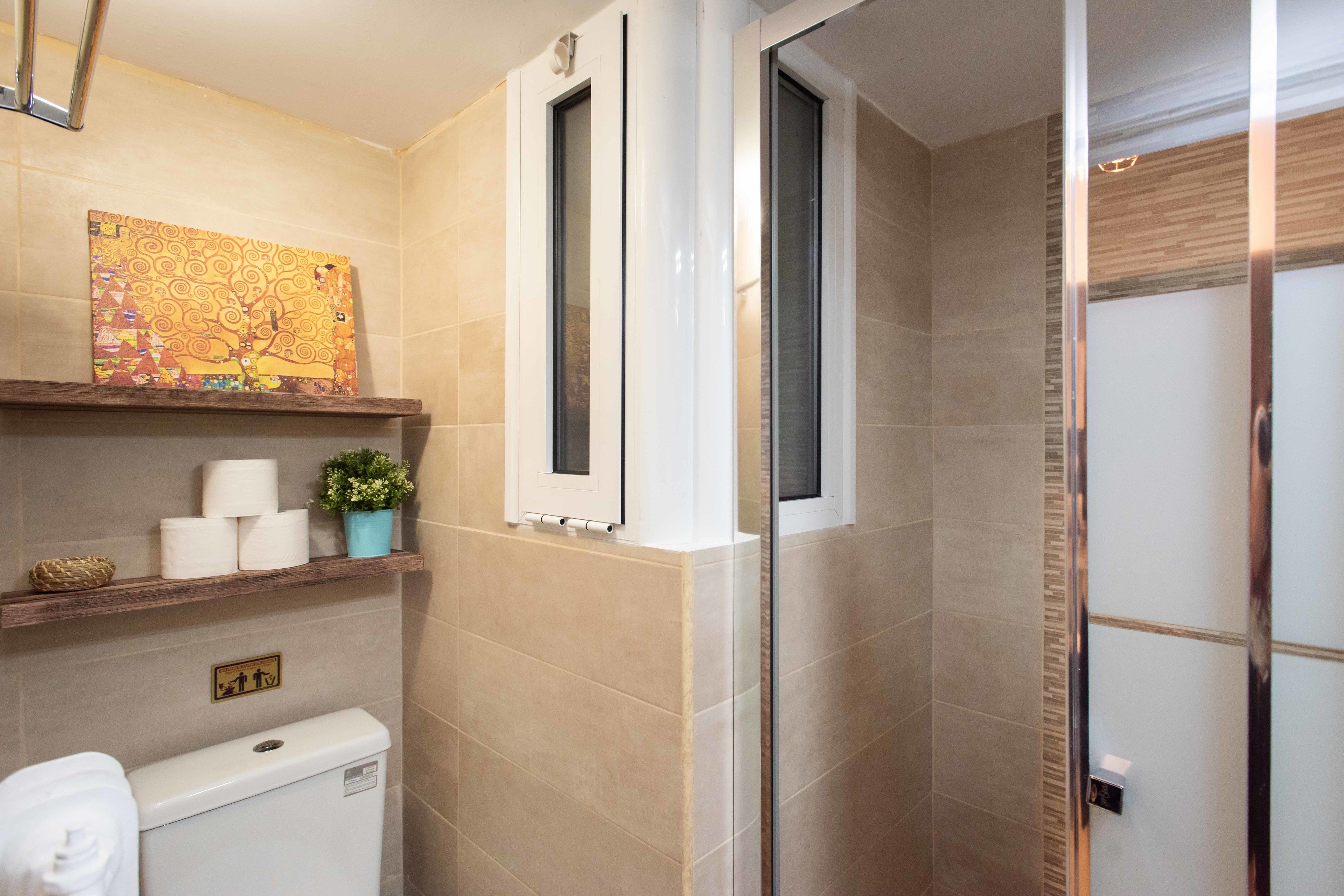 A bathroom with beige tiles, a toilet, a shower with a glass door, a shelf with toiletries, and a painting on the wall.