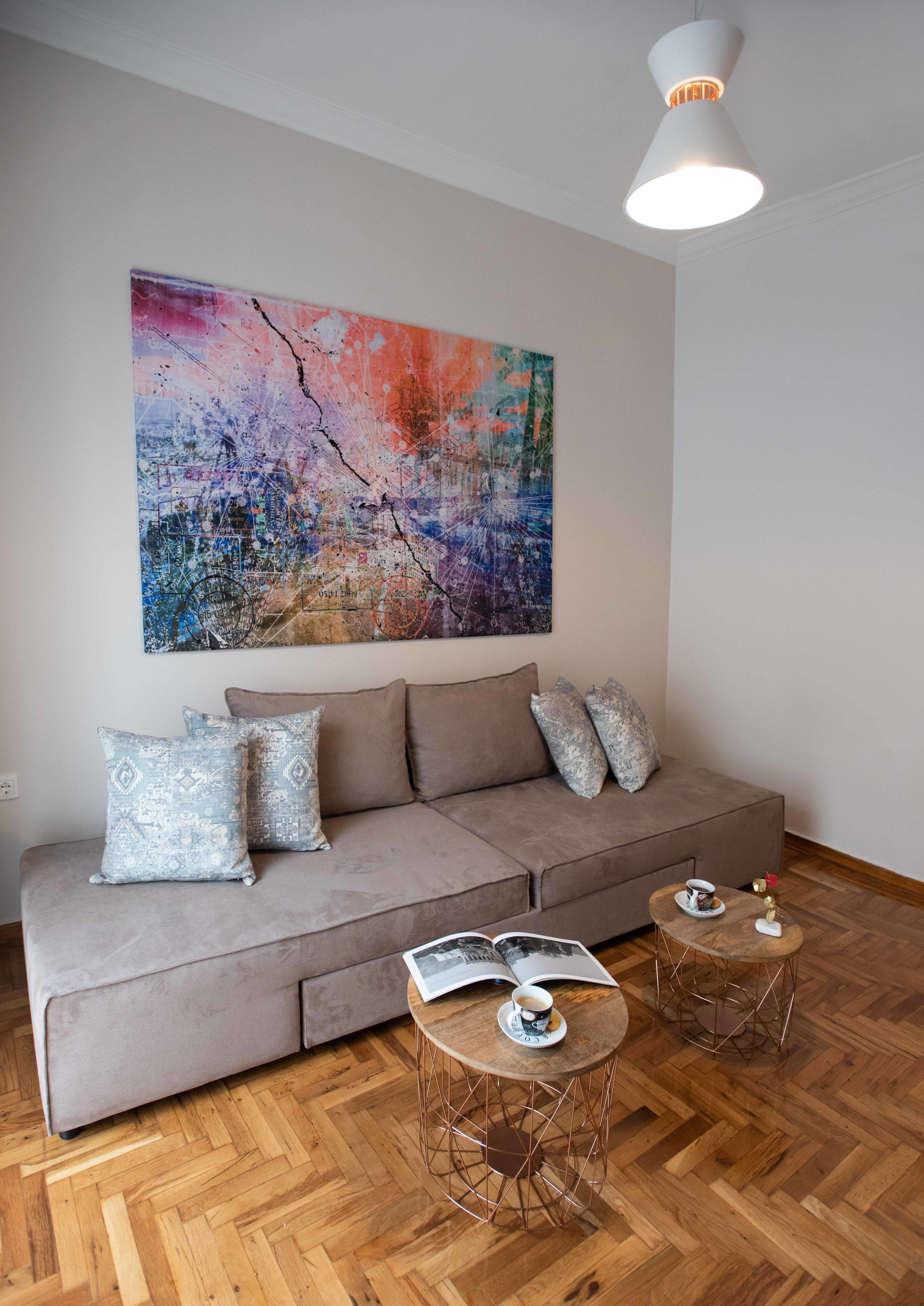 A modern living room with a grey sofa, patterned cushions, a large abstract painting, and two round coffee tables with books and a cup.