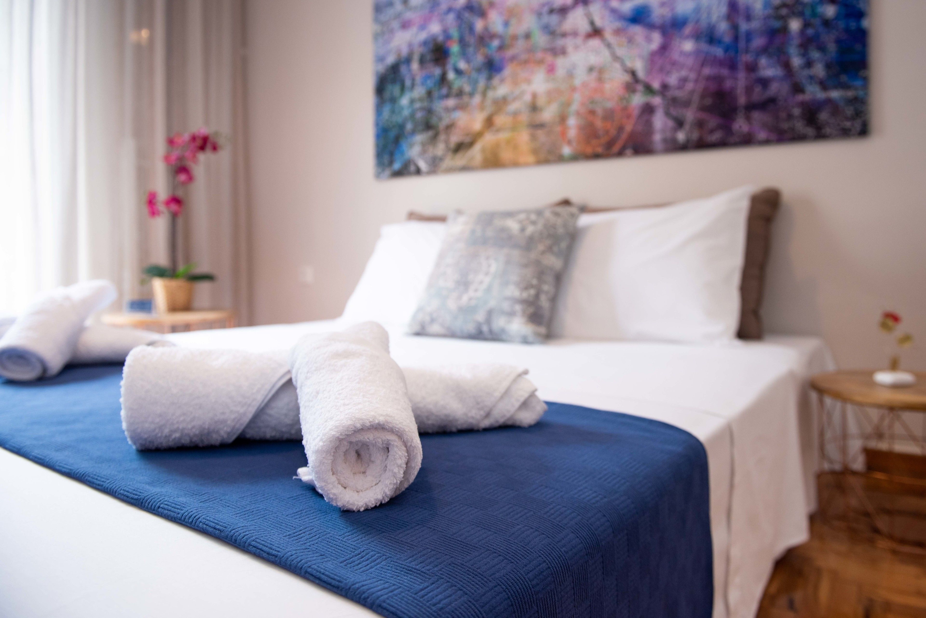 Een net opgemaakt bed met een blauwe spreideken en opgerolde witte handdoeken, een groot abstract schilderij aan de muur, en een orchidee in een pot op een bijzettafeltje.