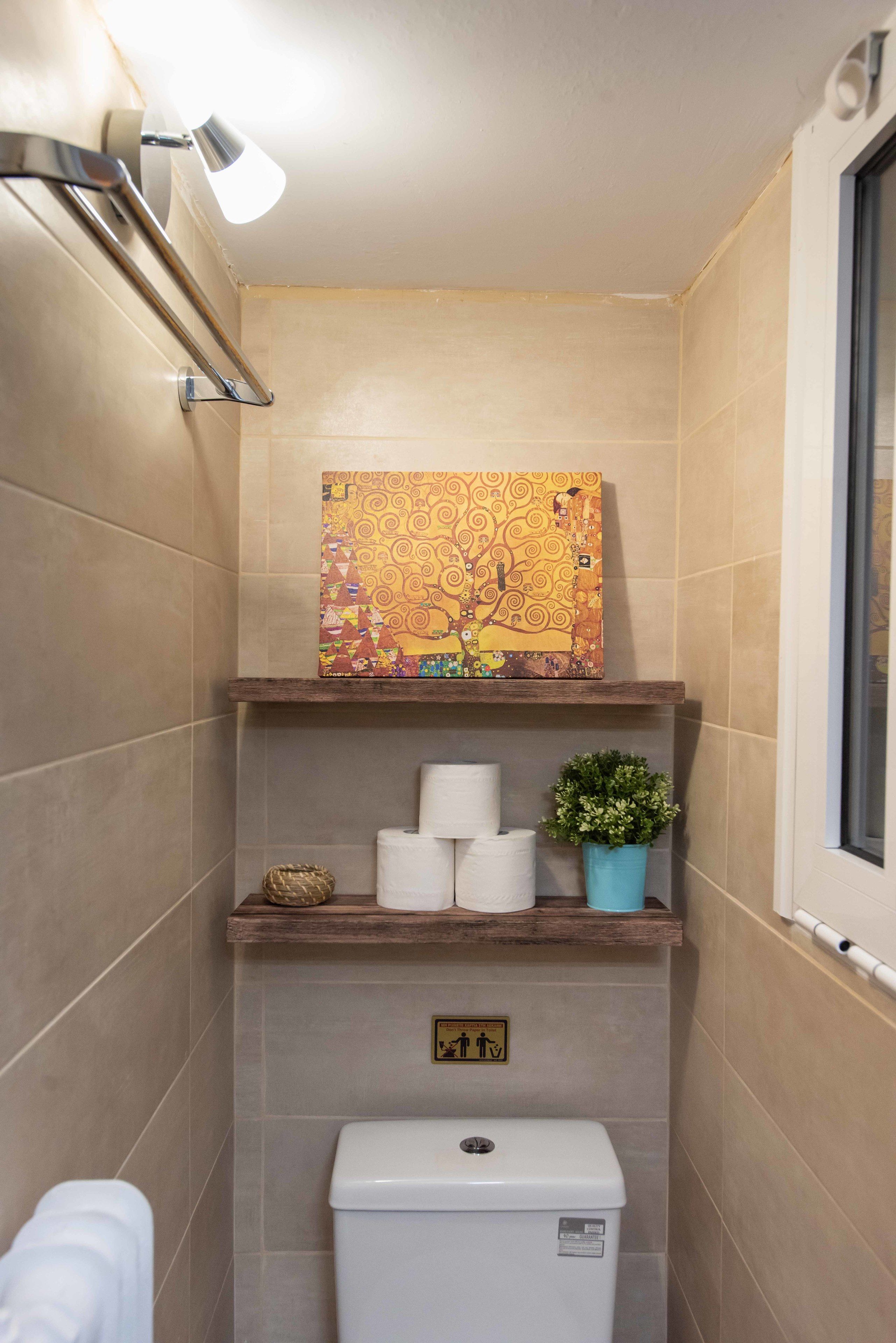Un baño con baldosas color crema, un inodoro, una repisa con papel higiénico y una planta en maceta, y un cuadro sobre la repisa.