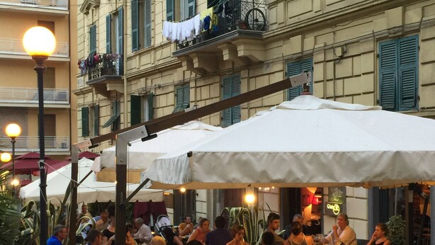 Un espace extérieur aménagé pour les repas, avec plusieurs tables et des parasols, entouré d’immeubles dotés de balcons et de fenêtres.