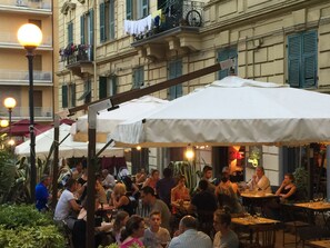 An outdoor dining area with multiple tables and umbrellas, surrounded by buildings with balconies and windows. - Echi del Mare (La Spezia)