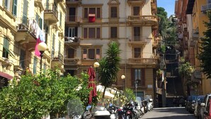 Une rue étroite bordée de motos et de voitures garées, des immeubles dotés de balcons et un ciel dégagé.