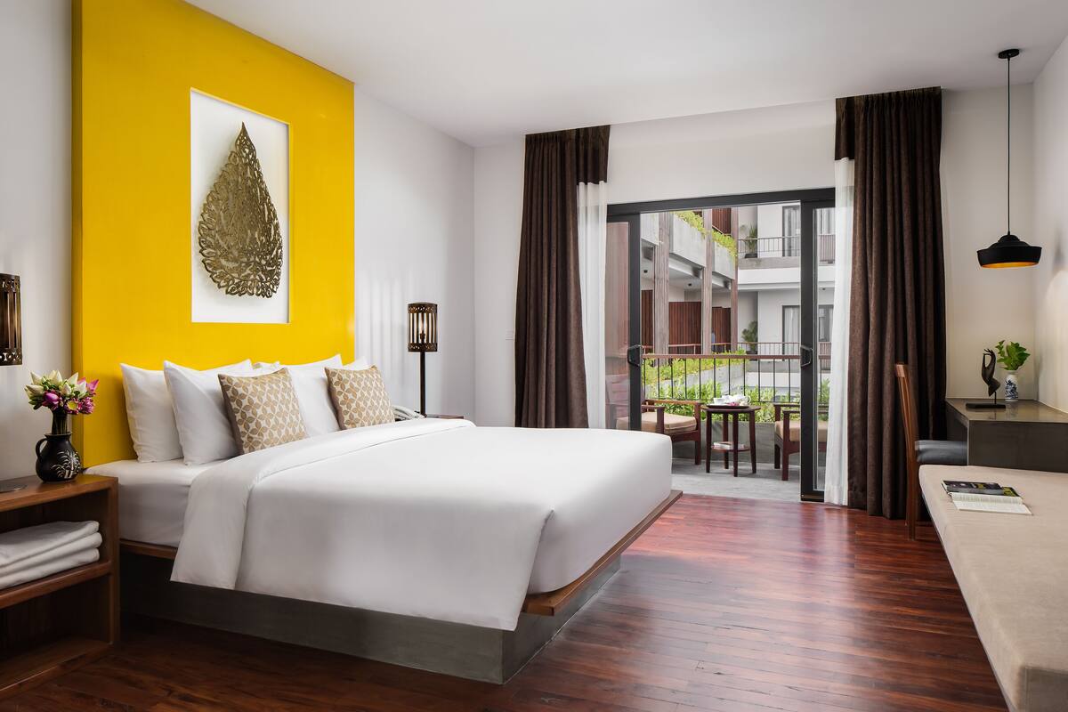 Une chambre d’hôtel moderne avec un grand lit, un balcon offrant une vue sur l’immeuble et un mur d’accent jaune.