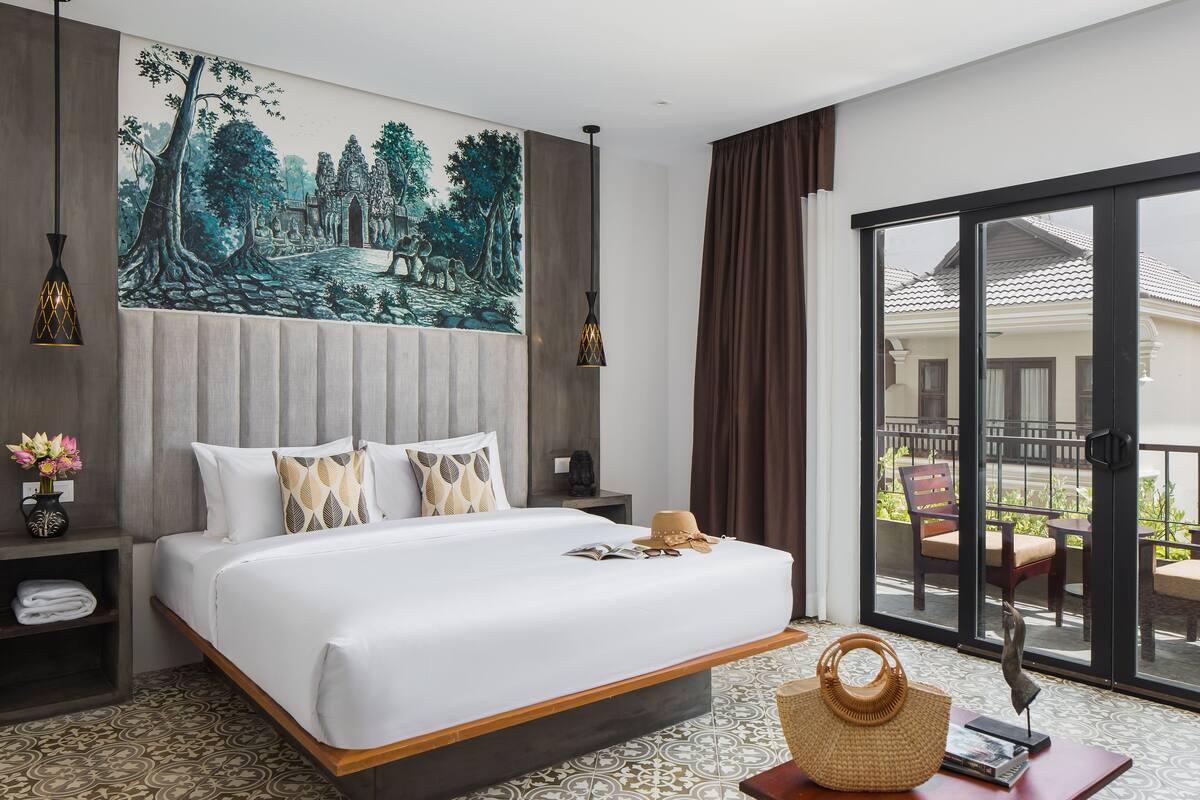 Une chambre d’hôtel avec un grand lit, un tableau au mur, un balcon avec des chaises et une vue sur un immeuble.