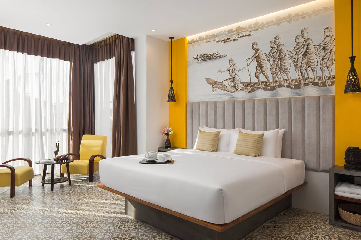 Une chambre d’hôtel avec un grand lit, une chaise, une petite table avec un plateau, et une fresque murale.