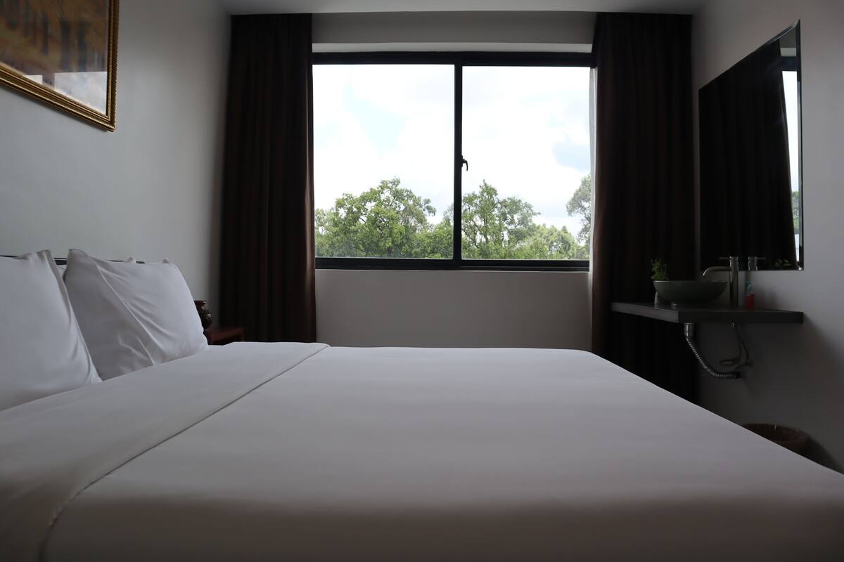Une chambre d’hôtel avec un grand lit, une fenêtre avec des rideaux, une table de chevet avec un bol et une bouteille, et un miroir au mur.
