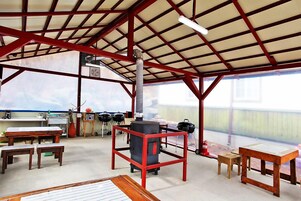 Un espacio al aire libre techado con techo metálico, mesas, bancos y equipo para parrillada.