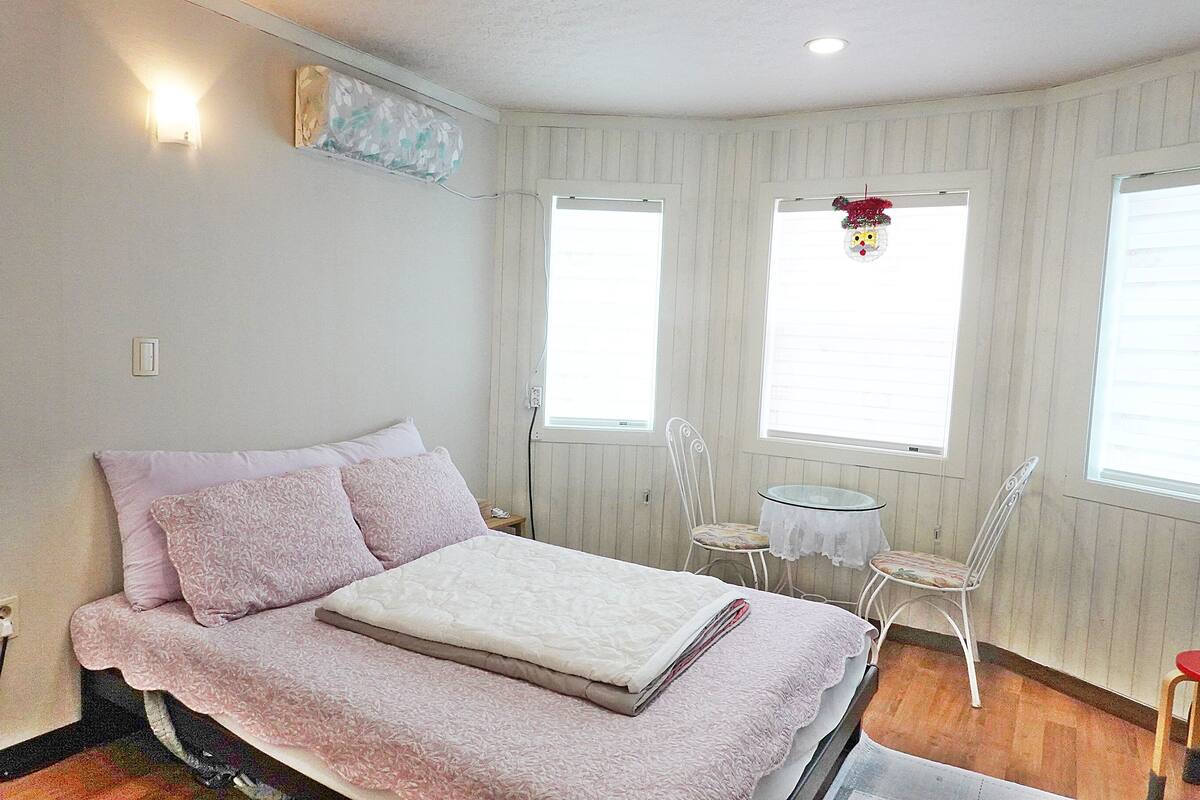 A bedroom with a bed, two chairs, a small table, and two windows with blinds.