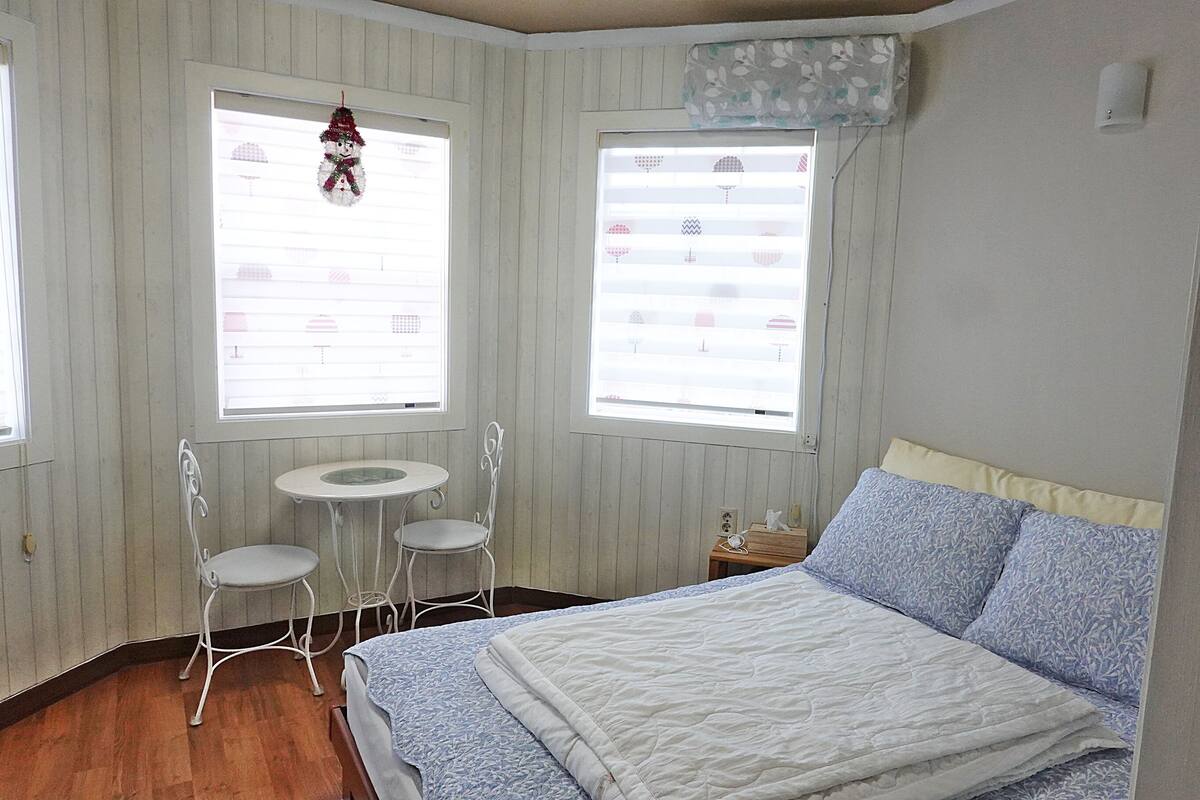 A bedroom with a bed, two white chairs, a small table, and two windows with blinds.
