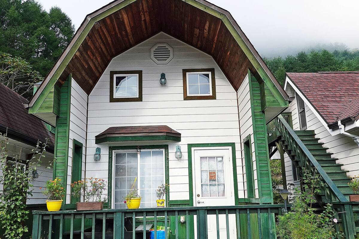 A white house with green trim and a porch, surrounded by greenery.