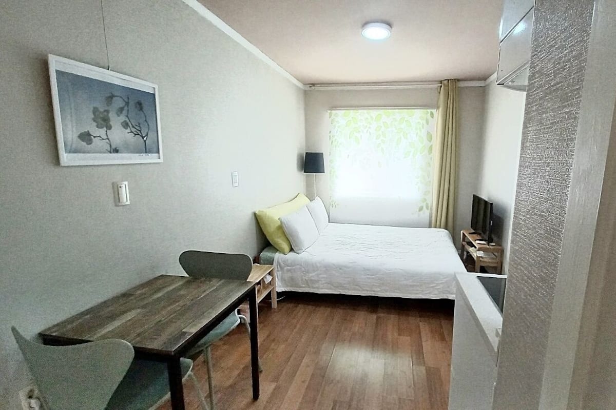A small, well-lit room with a bed, a desk, and a chair. There is a painting on the wall and a window with curtains.