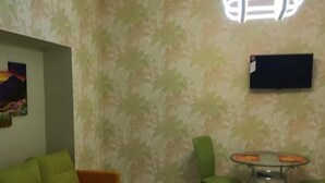 Huoneessa on sohva, pöytä tuoleineen, seinälle asennettu televisio ja kuviollinen tapetti.