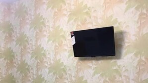 Huoneessa on seinälle asennettu taulutelevisio, kaksi vihreää tuolia ja lasitasoinen pöytä, jonka päällä on alusta.