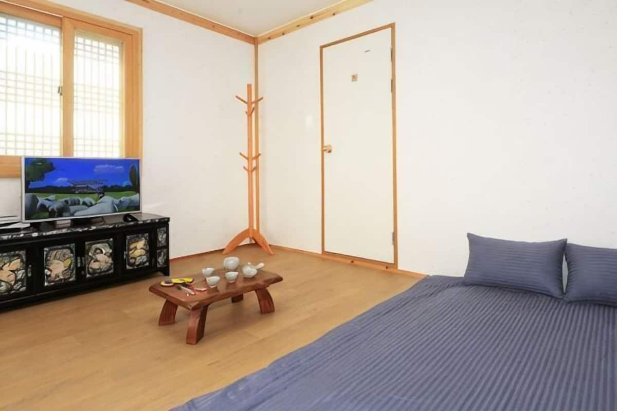 A room with a bed, a TV, a wooden stand with items, a coat rack, and a door.