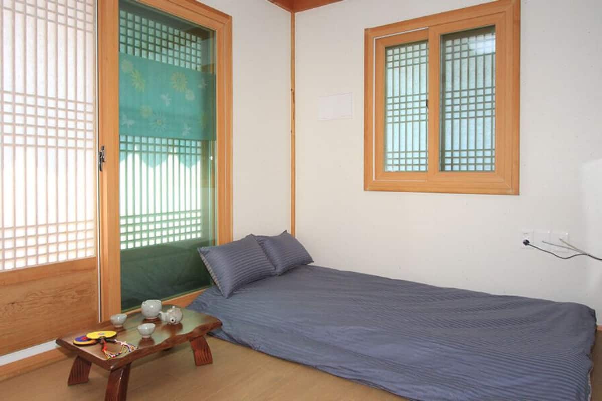 A small room with a single bed, a wooden table with a cup and a teapot, and a window with a grid pattern.