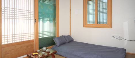 Una habitación pequeña con una cama individual, una mesa de madera con una taza y una tetera, y una ventana con diseño de cuadrícula.