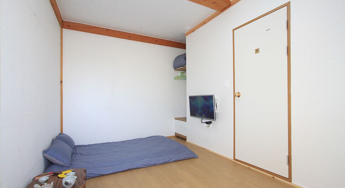 A small, sparsely furnished room with a single bed, a television mounted on the wall, and a wooden door.