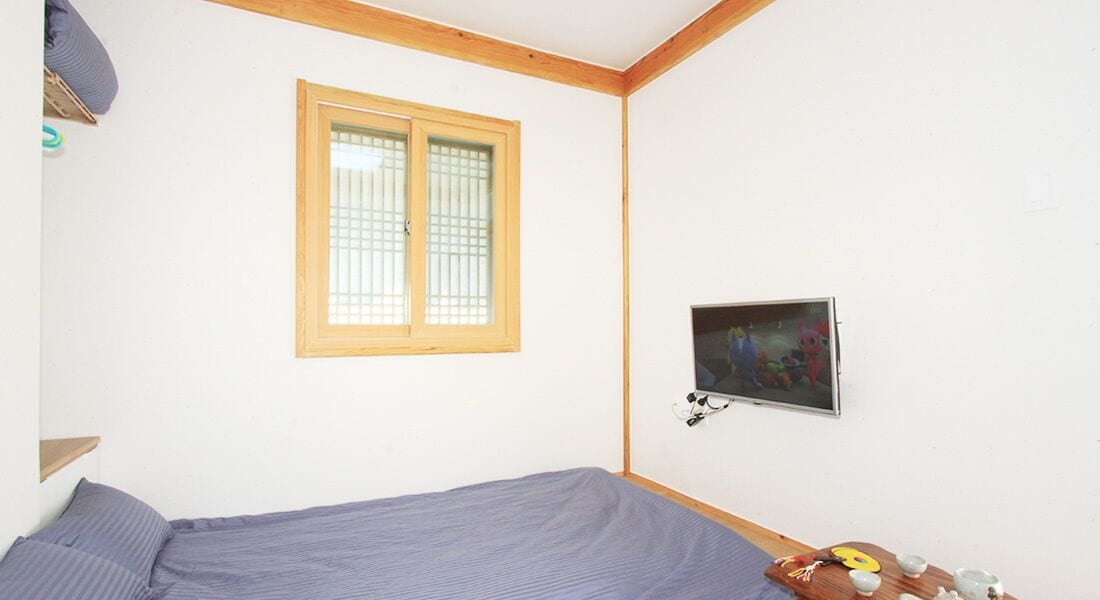 A bedroom with a single bed, a window with wooden frames, a wall-mounted TV, and a small table with a teapot and cups.