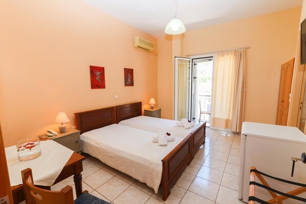 Habitación de hotel con cama doble, un escritorio con silla, una mesita con lámpara, un refrigerador y una puerta corrediza de vidrio al balcón.