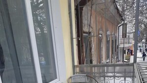 A balcony with a glass table and two chairs, a yellow wall, and a metal railing.