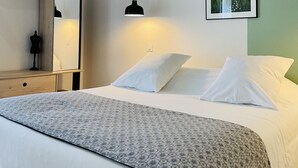 Un lit bien fait, avec du linge de lit blanc et un couvre-lit à motifs, une lampe de chevet, un climatiseur fixé au mur et un tableau représentant un paysage.