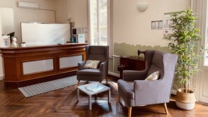 Un espace d’accueil d’hôtel avec un comptoir en bois, deux fauteuils, une petite table et une plante en pot.