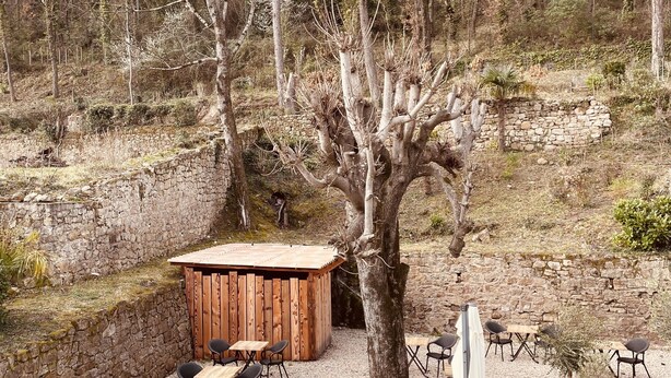 Un espace patio agrémenté de murs en pierre, de tables et de chaises en bois, entouré d’arbres et d’une pelouse verdoyante.