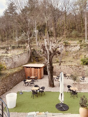 Un patio con paredes de piedra, mesas y sillas de madera, rodeado de árboles y un césped.