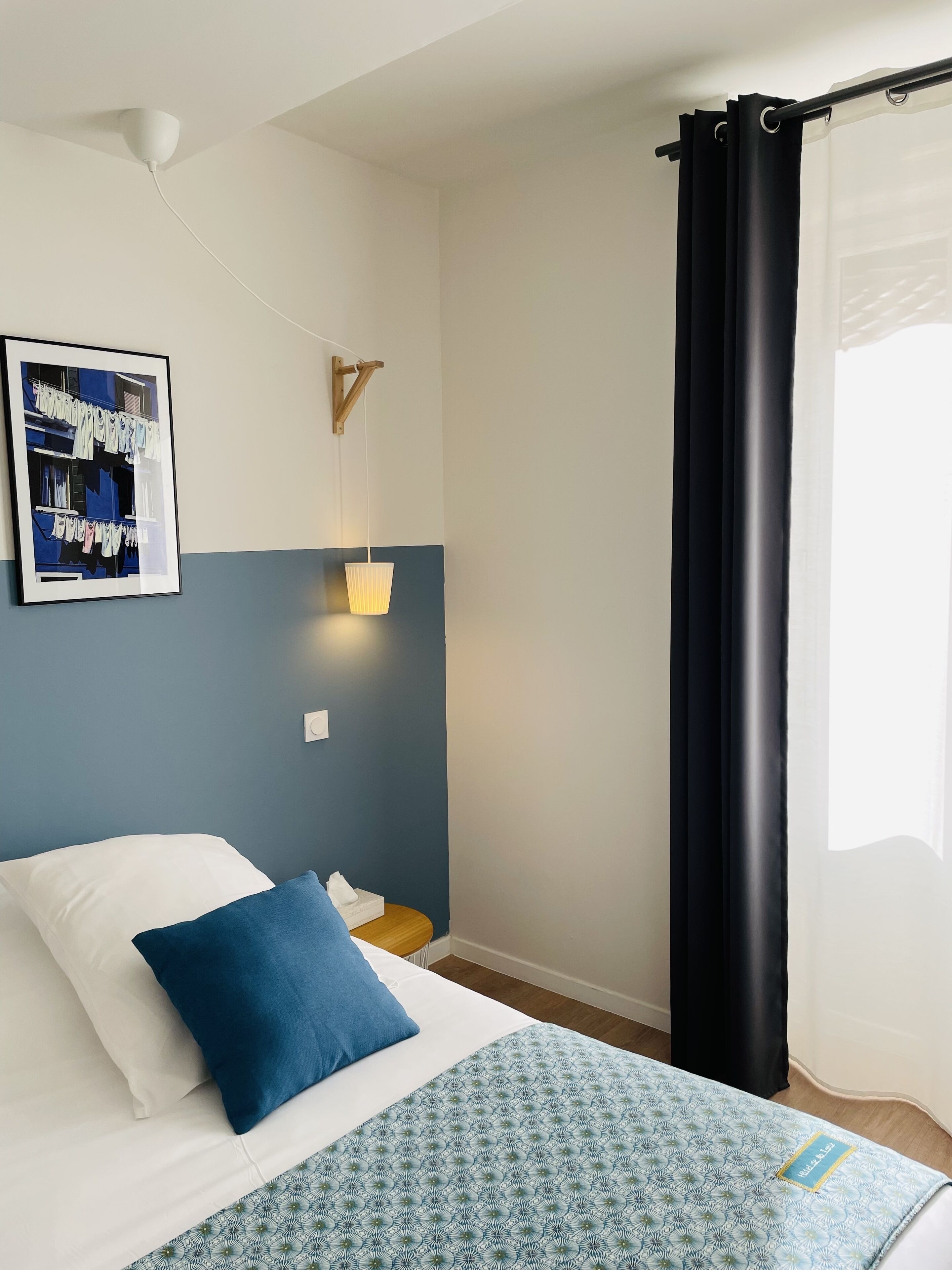 Une chambre à coucher avec un lit, des oreillers, une lampe fixée au mur, un tableau encadré et un rideau.