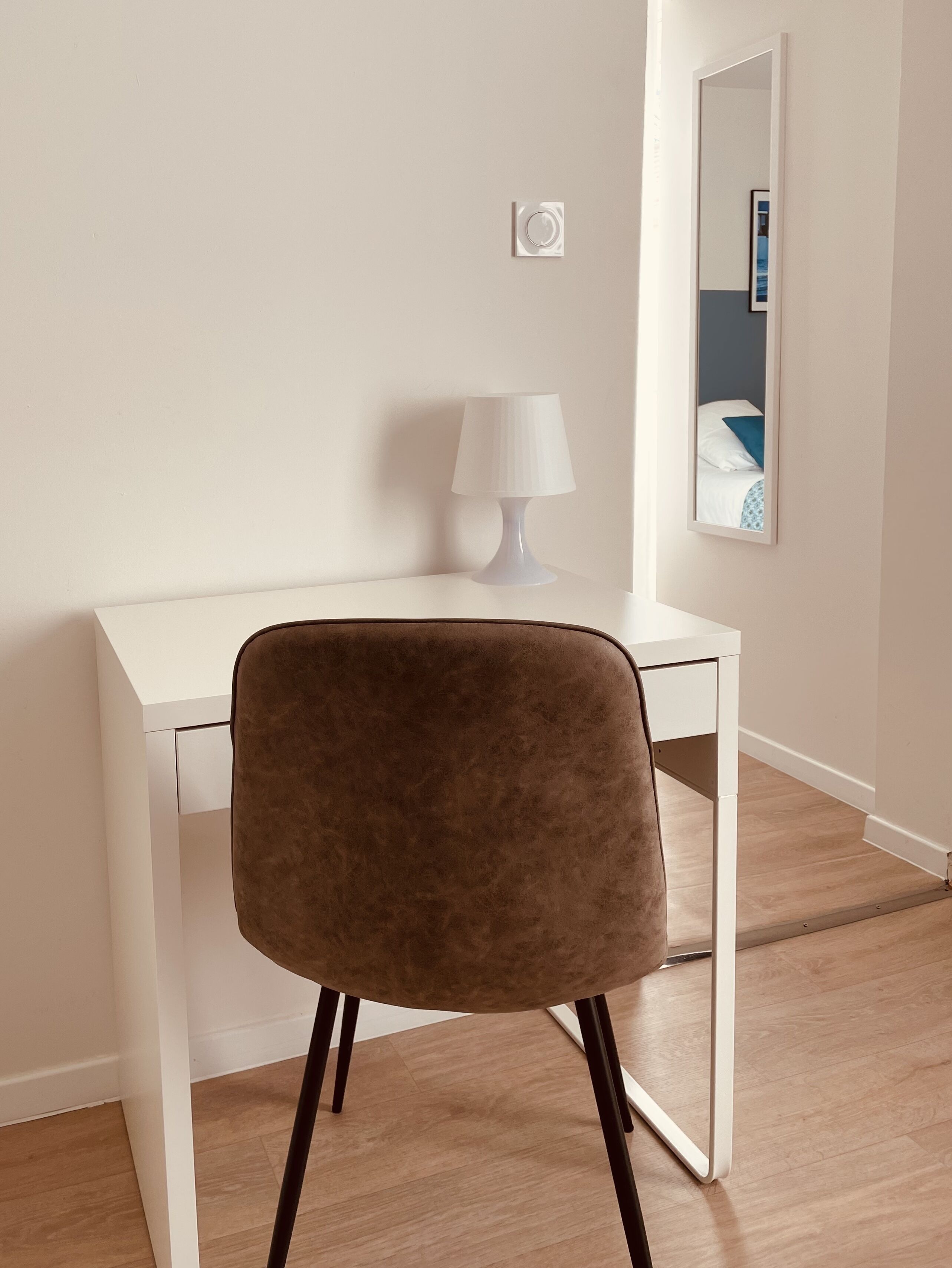 A modern room with a white desk, a brown chair, a white lamp, and a mirror on the wall.