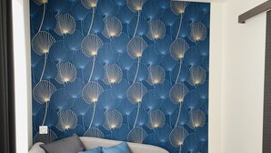 Un salon moderne avec un canapé gris, des coussins bleus, une table basse ronde en bois et un papier peint bleu orné d’un motif de coquillages.