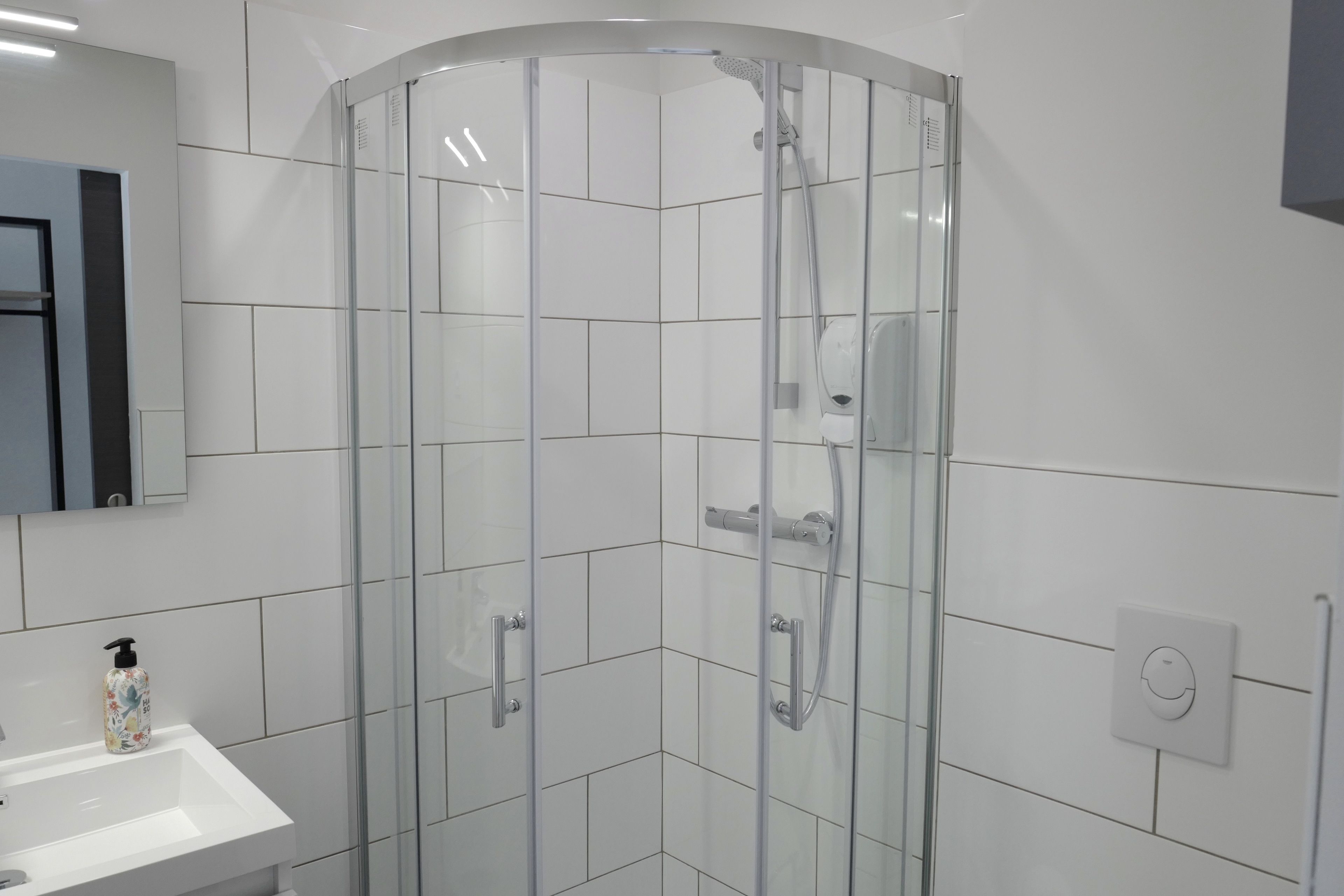 A modern bathroom with a glass shower enclosure, white tiles, a sink with a soap dispenser, and a mirror.