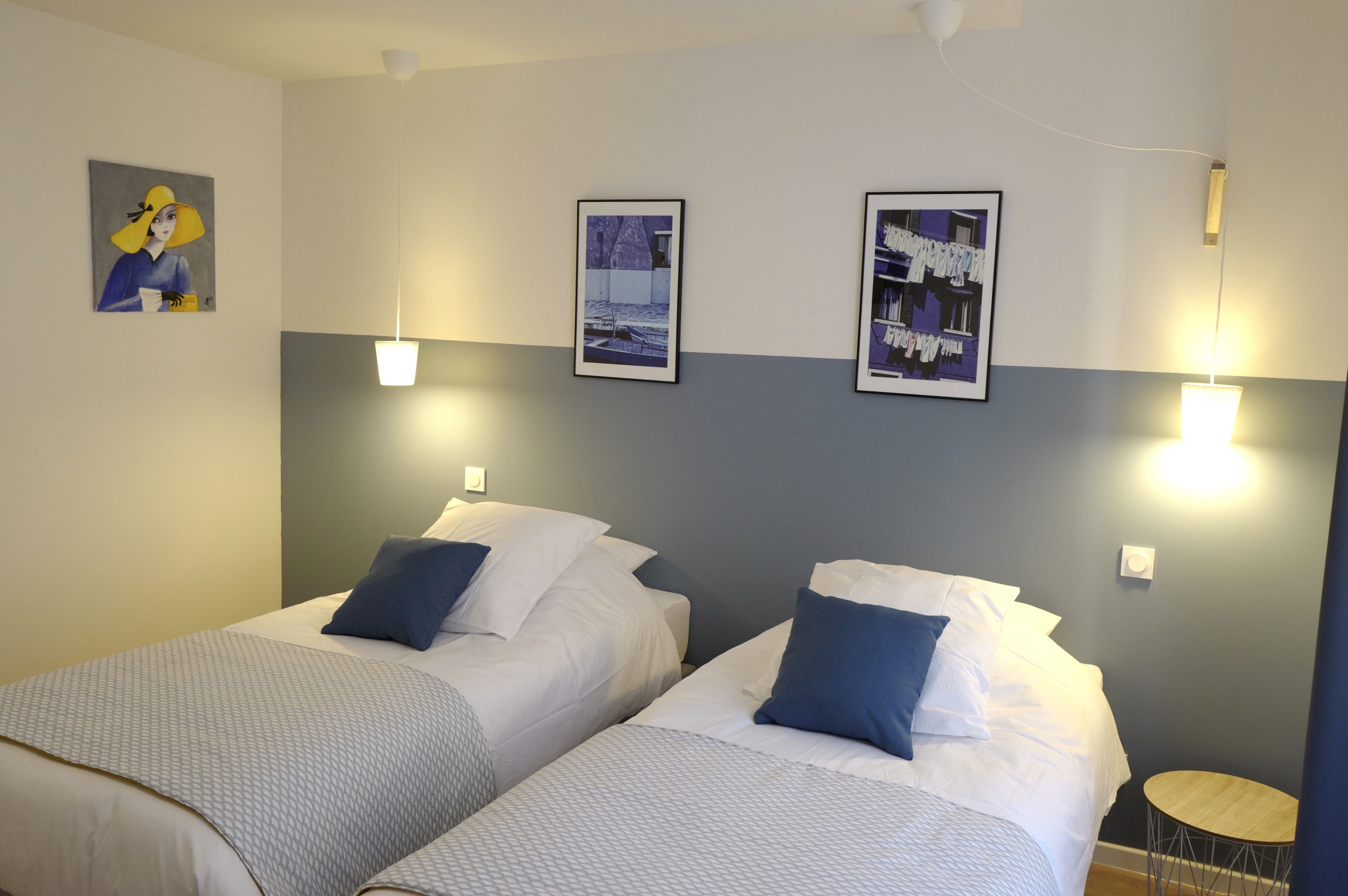 A hotel room with two beds, each with white linens and blue pillows, a small round wooden table, and wall-mounted light fixtures.